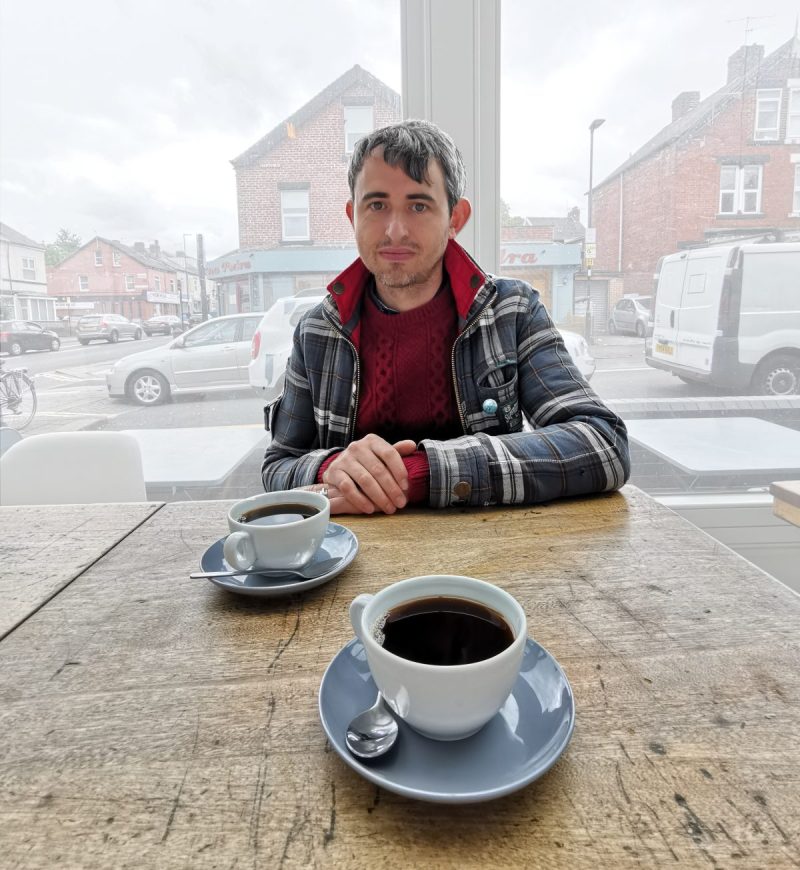 Dan sitting at a table in a window with two cups of coffee in front of him