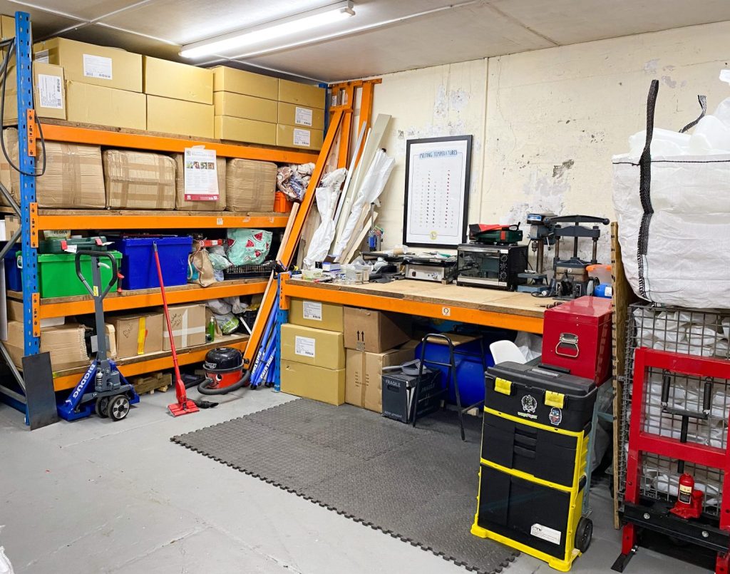 A photo of a workshop with a cream wall and an orange framed work bench and racking. There are boxes on the shelves, matting on the floor and a Henry hoover alongside the work bench.